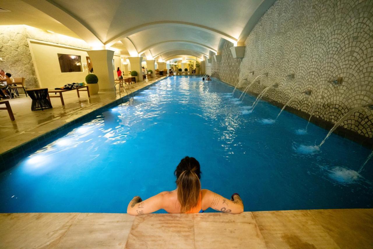Buona Vitta Gramado Resort & Spa By Gramado Parks Exteriör bild The photo shows an indoor swimming pool with a long, narrow design. The water is a clear blue, and there are features such as fountains along one side. In the foreground, a person with long hair is sitting on the edge of the pool, looking out over th