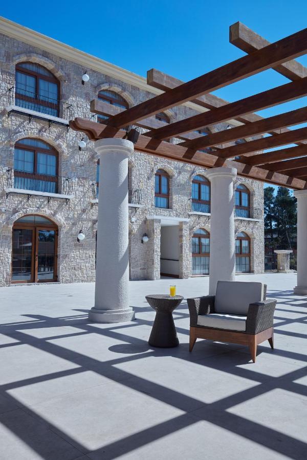 Buona Vitta Gramado Resort & Spa By Gramado Parks Exteriör bild The photo shows a well-designed outdoor space with a stone wall building in the background. There are several tall columns that support a wooden pergola overhead, creating a shaded area. In this space, there is a stylish chair and a small table besid