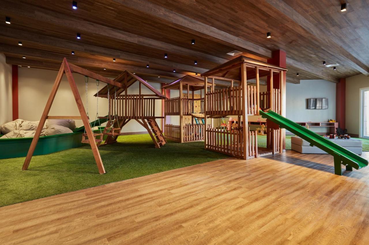 Buona Vitta Gramado Resort & Spa By Gramado Parks Exteriör bild The photo shows an indoor play area designed for children. It features a wooden play structure that includes multiple levels, slides, and a swing set. The floor is covered with wooden planks, and there is a green carpet-like area that adds a soft tou