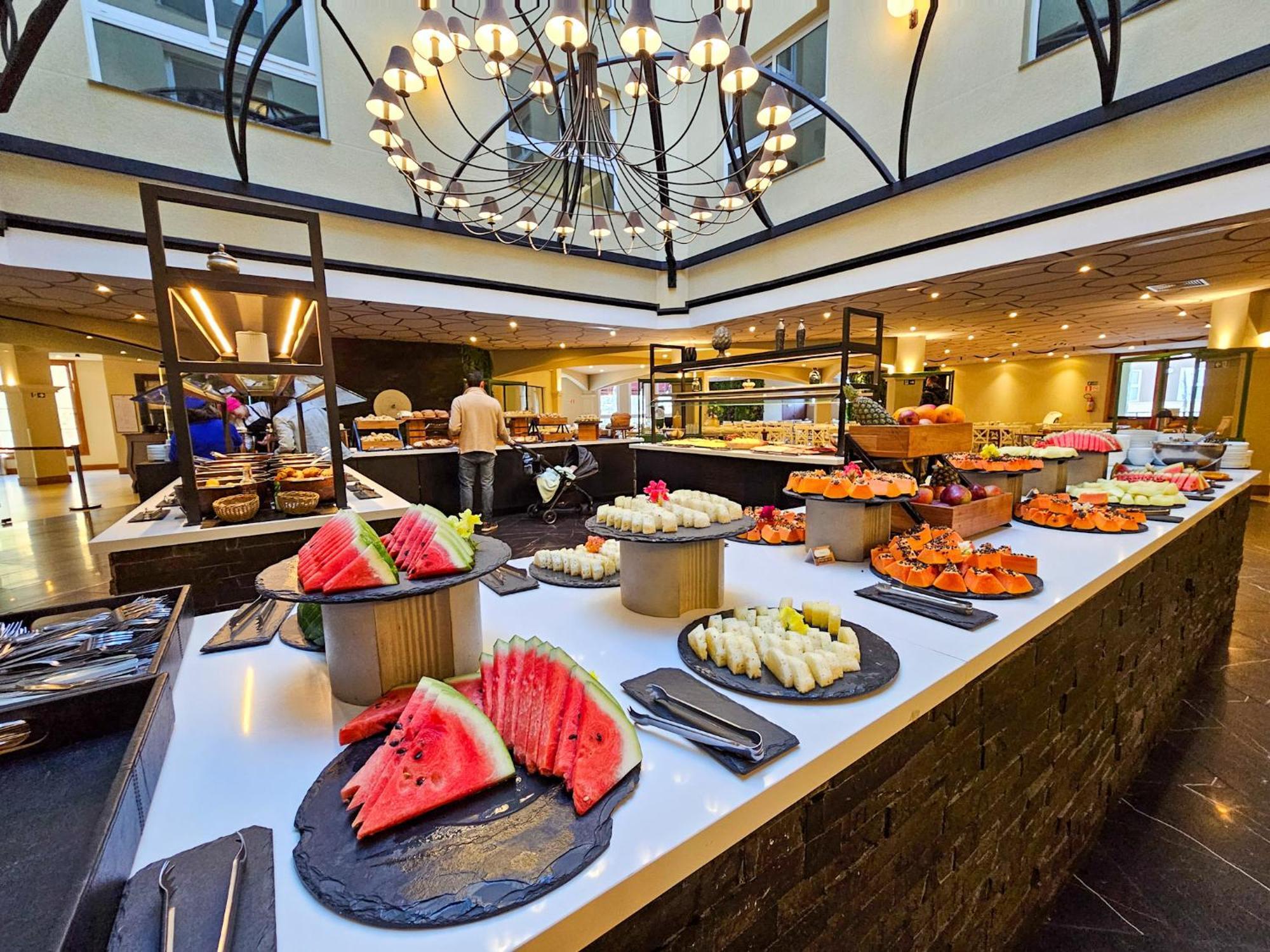 Buona Vitta Gramado Resort & Spa By Gramado Parks Exteriör bild The photo depicts a lavish buffet setup in a dining area. In the foreground, there are several platters featuring an assortment of colorful food items, primarily fruits like watermelon and various types of sushi or rolls. The buffet table is elegantl