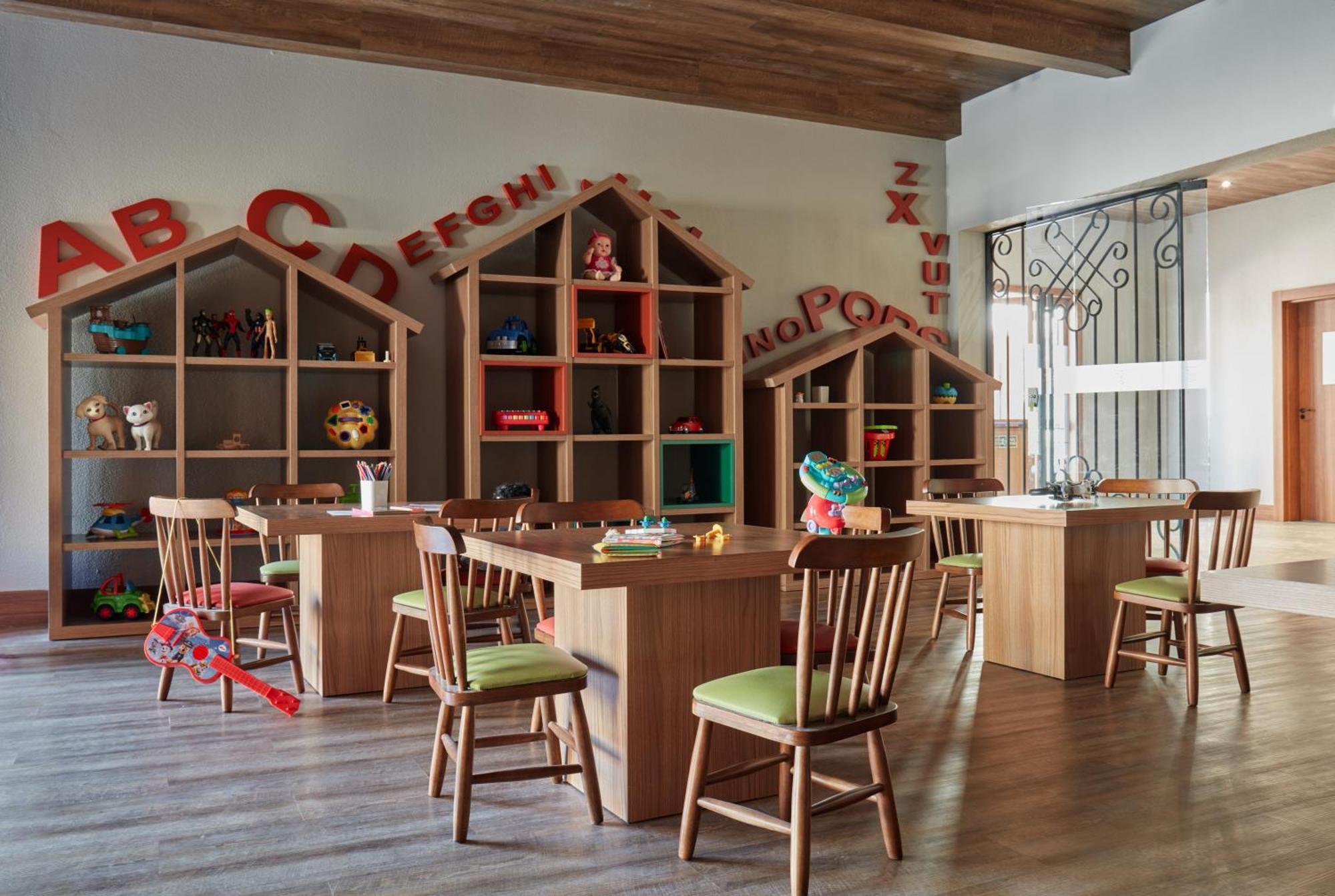 Buona Vitta Gramado Resort & Spa By Gramado Parks Exteriör bild The photo shows a classroom or playroom designed for children. It features wooden furniture, including several tables and chairs with green cushions. In the background, there are large, open shelving units shaped like houses, displaying various toys 