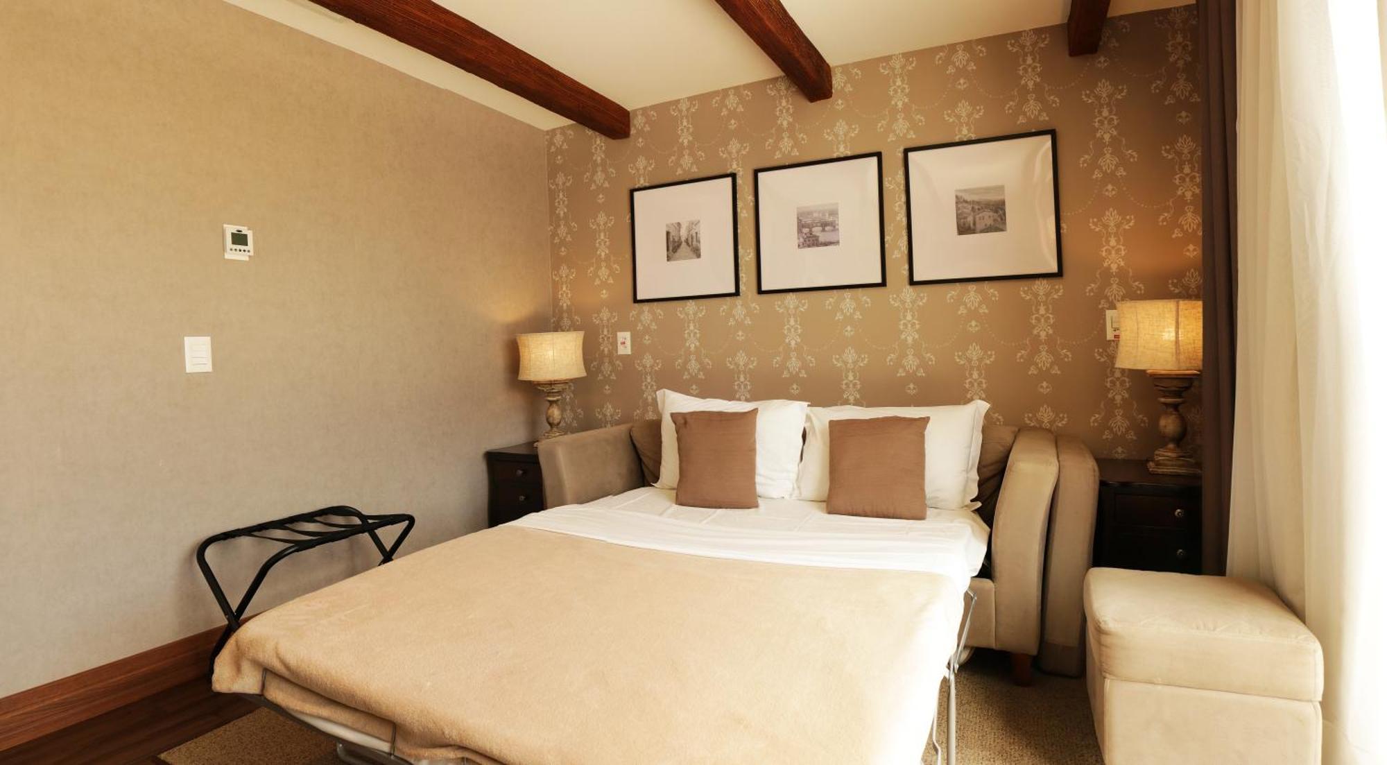 Buona Vitta Gramado Resort & Spa By Gramado Parks Exteriör bild The photo shows a cozy bedroom with a neutral color palette. There is a bed with white linens and two brown decorative pillows. On either side of the bed, there are bedside tables with lamps. The walls feature a subtle floral pattern, and three frame