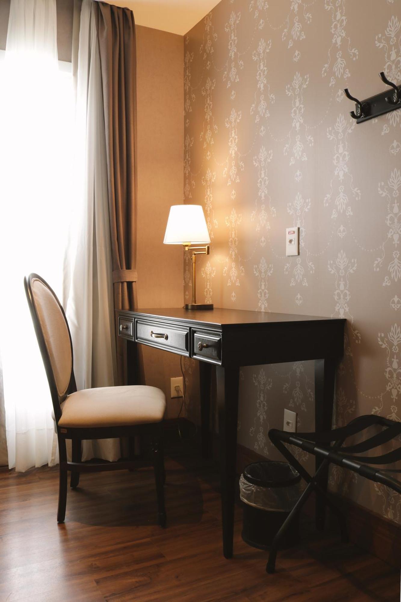 Buona Vitta Gramado Resort & Spa By Gramado Parks Exteriör bild The photo shows a small desk set against a wall with a decorative pattern. The desk is black with a few drawers and has a small lamp on it. Next to the desk, there is a chair with a light-colored cushion. Soft light is coming through a window, castin