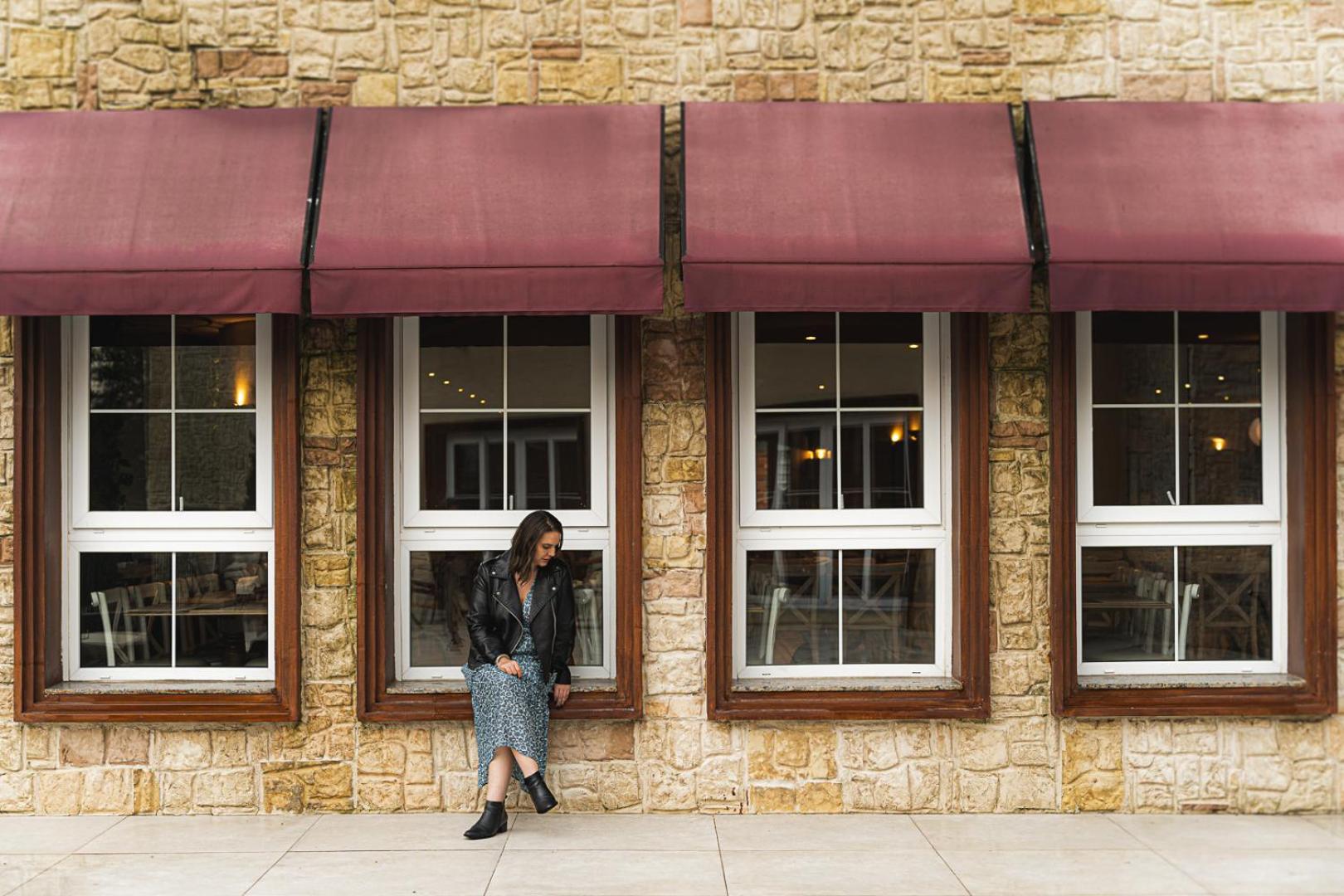 Buona Vitta Gramado Resort & Spa By Gramado Parks Exteriör bild The photo shows a woman standing against a stone wall with three large windows in front of her, each framed with wooden borders. The windows have maroon awnings above them. The woman is dressed in a long skirt and leather jacket, leaning slightly aga